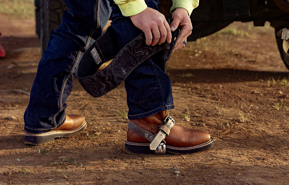 Close-up of a lineman, Dylan Edgmon, wearing brown leather work boots and attaching climbing gear while standing on dirt.